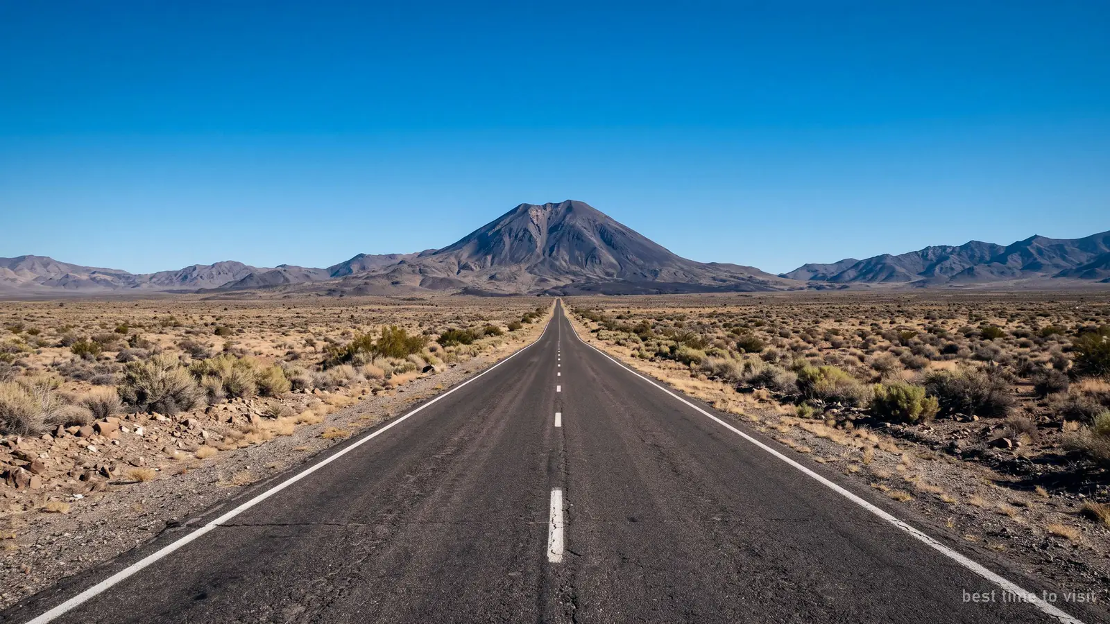 An empty desert road running towards a distant volcano under a clear sky