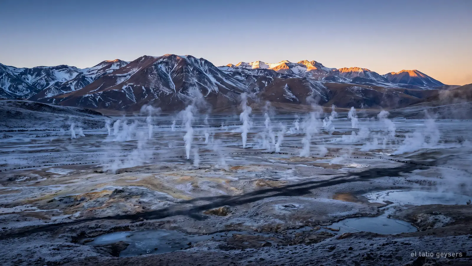 Steam venting from a high desert basin at first light, with snow-streaked mountains behind